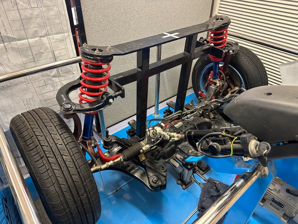 A car's front suspension and steering training rig on a stand: coil-over strut, control arms, knuckle, brake and wheel