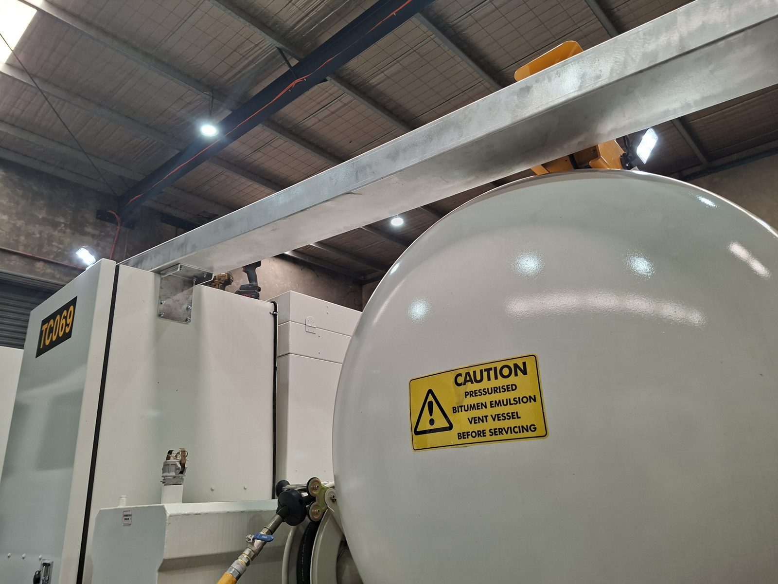 A white pressurised bitumen emulsion tank with a caution sticker and a hose reel